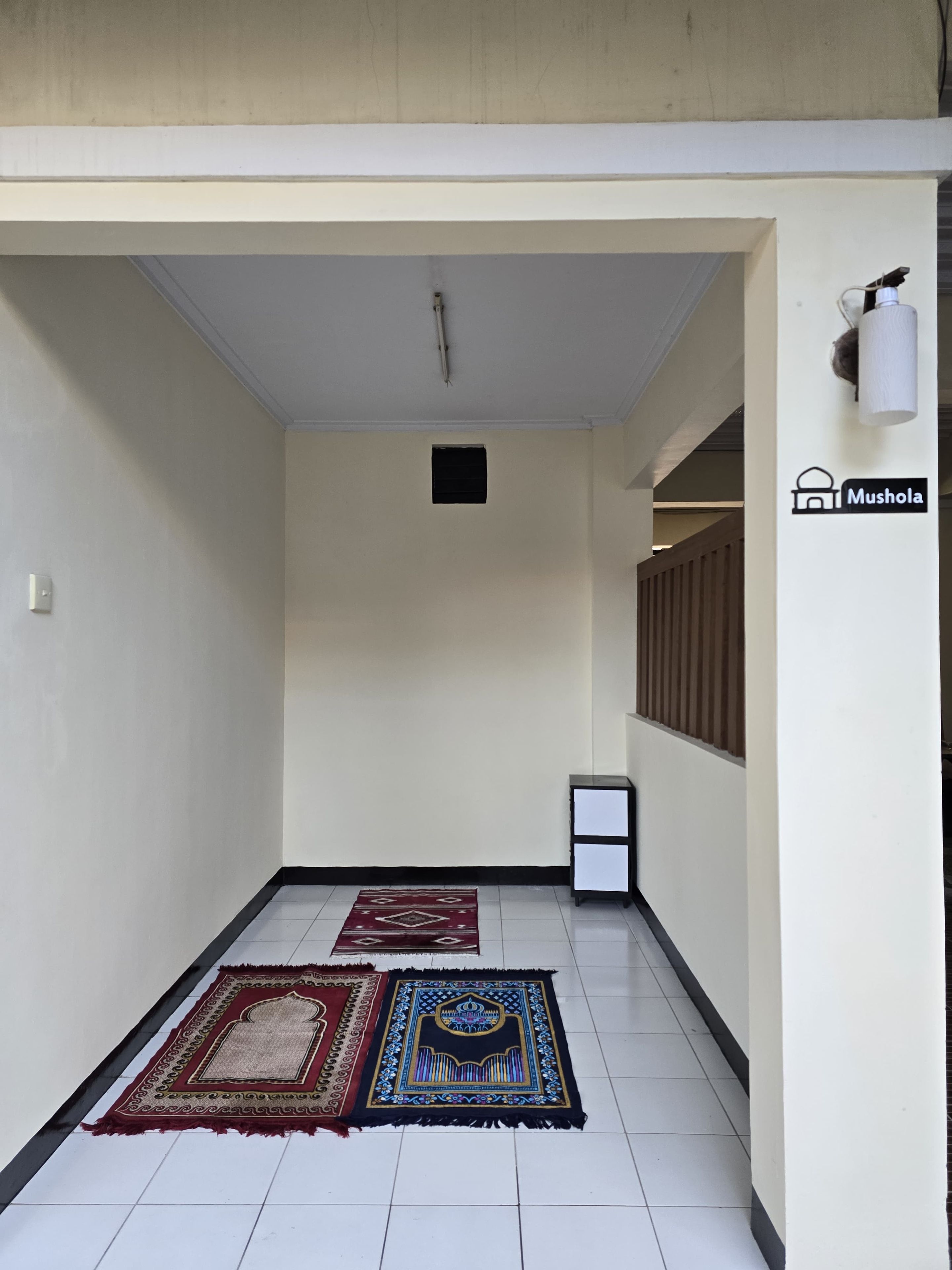 A hallway with two rugs, a wall-mounted light, and a sign for 'Mushola'.