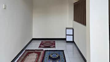 A hallway with two rugs, a wall-mounted light, and a sign for 'Mushola'.