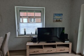 A living room with a television on a wooden stand, a window with blinds, a small table with decorative items, and a chair. - Holiday home with 2 bedrooms (Neßmersiel)