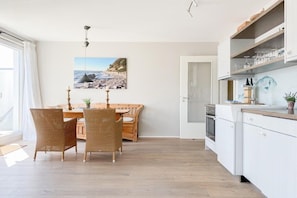 A kitchen with white cabinets, a dining area with wicker chairs, and a painting of a beach scene. - Holiday apartment with 1 bedroom (Timmendorfer Strand)