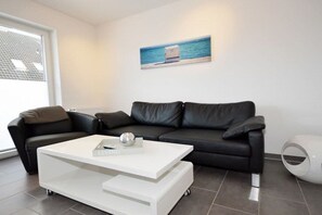A modern living room with a black leather sofa, a white coffee table, and a painting of a beach scene on the wall. - Holiday apartment with 1 bedroom (Norddeich)