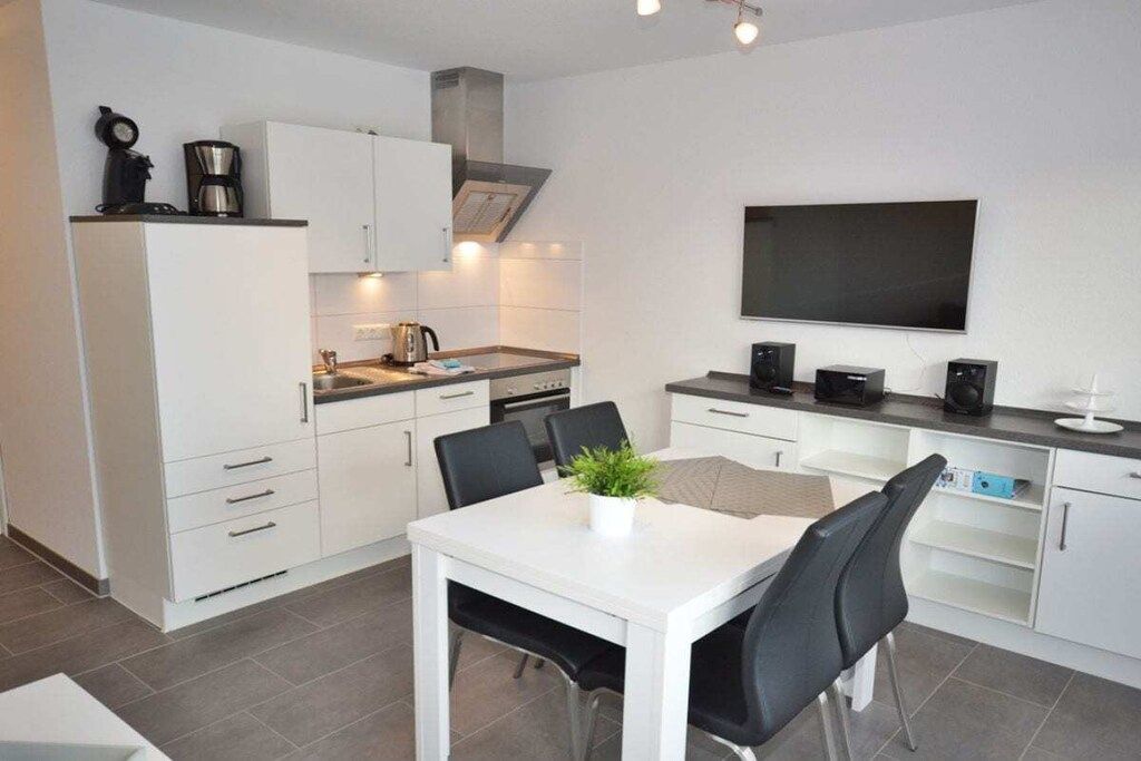 A modern kitchen with white cabinetry, a dining table with chairs, a flat-screen TV mounted on the wall, and a coffee maker on the countertop.