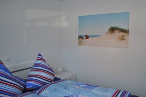 A bedroom with a bed, striped pillows, a nightstand, a lamp, and a beach-themed wall hanging. - Holiday apartment with 1 bedroom (Norddeich)