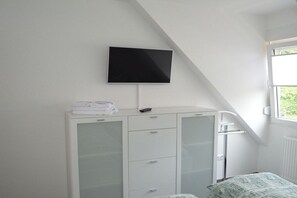 Een slaapkamer met een bed, een witte kledingkast, een aan de muur bevestigde televisie en een raam dat voor natuurlijk licht zorgt.