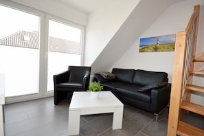 A modern living room with a black leather sofa, a white coffee table, and a painting of a lighthouse on the wall. - Holiday apartment with 1 bedroom (Norddeich)
