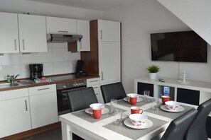 A compact kitchen with white cabinets, a stove, and a dining area with a table set for four. - Holiday apartment with 1 bedroom (Norddeich)