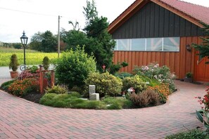 A building with a brick pathway leading to a wooden door, surrounded by a landscaped garden with various plants and a stone structure. - Living at the Moor 2 (Ihlow-Westersander)