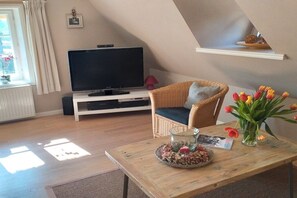 Ein gemütliches Wohnzimmer mit Fernseher, einem Korbstuhl, einem Couchtisch mit einer Vase voller Tulpen und einem kleinen Regal über dem Sessel.