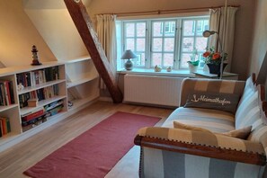 Une pièce chaleureuse avec un plafond en pente, une bibliothèque, une fenêtre avec des rideaux, un radiateur, un canapé avec un coussin et une lampe.