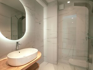 A modern bathroom with a round mirror, a white sink, and a glass-enclosed shower. - Natural Stone House 6 (Sitia)