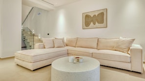 A modern living room with a large L-shaped sectional sofa, a round coffee table, and a staircase with glass railings. - Melas Cretan Horizon (Kefalas)