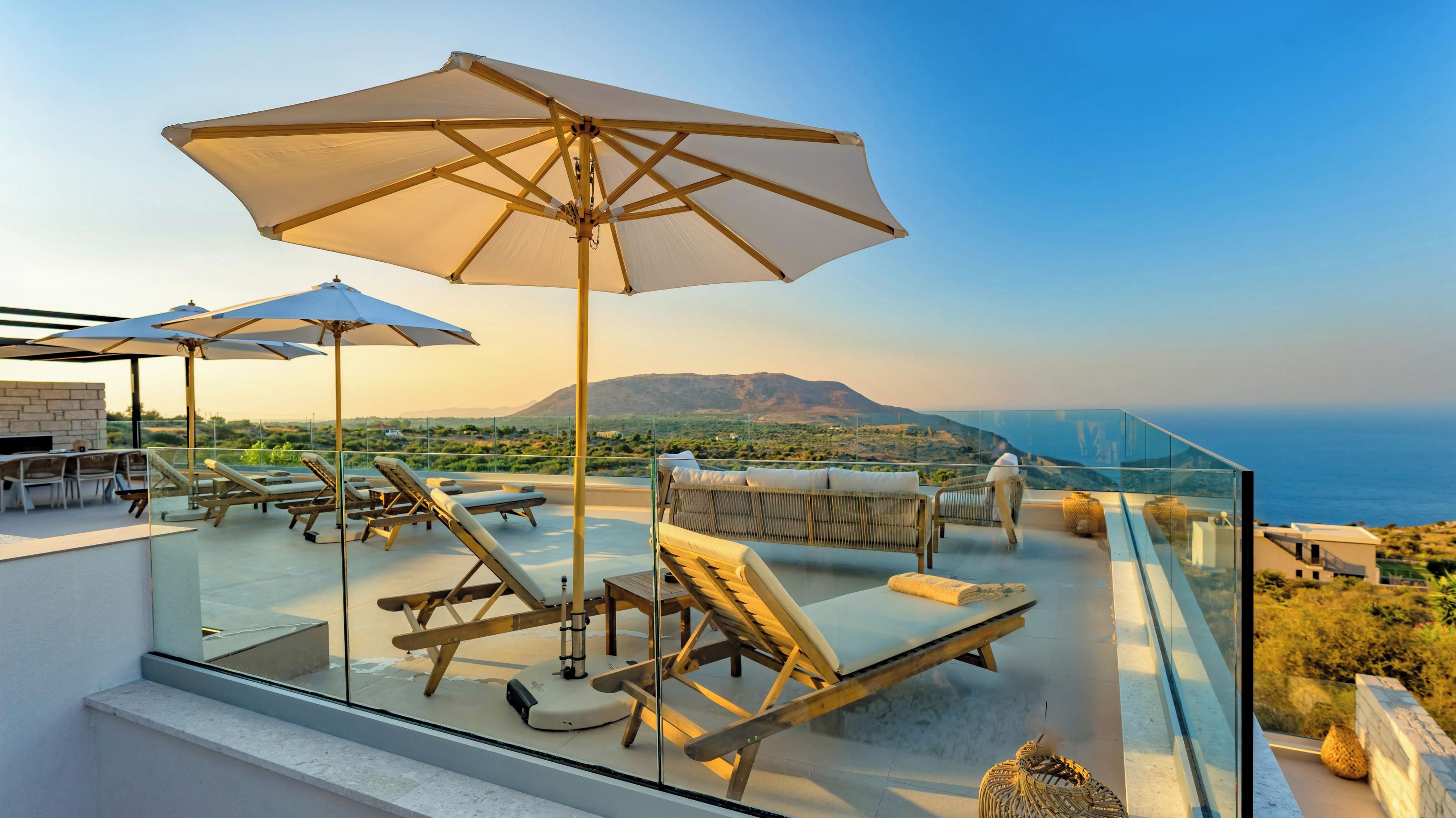 A rooftop terrace with lounge chairs, umbrellas, and a glass balustrade overlooking a landscape with mountains and the sea.
