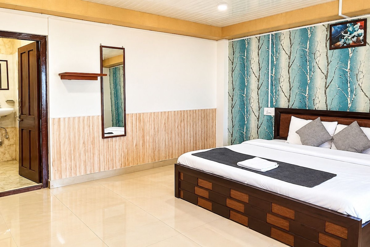 A hotel room with a large bed, a wooden headboard, a mirror, a bathroom with a sink, and a wall with a tree pattern.