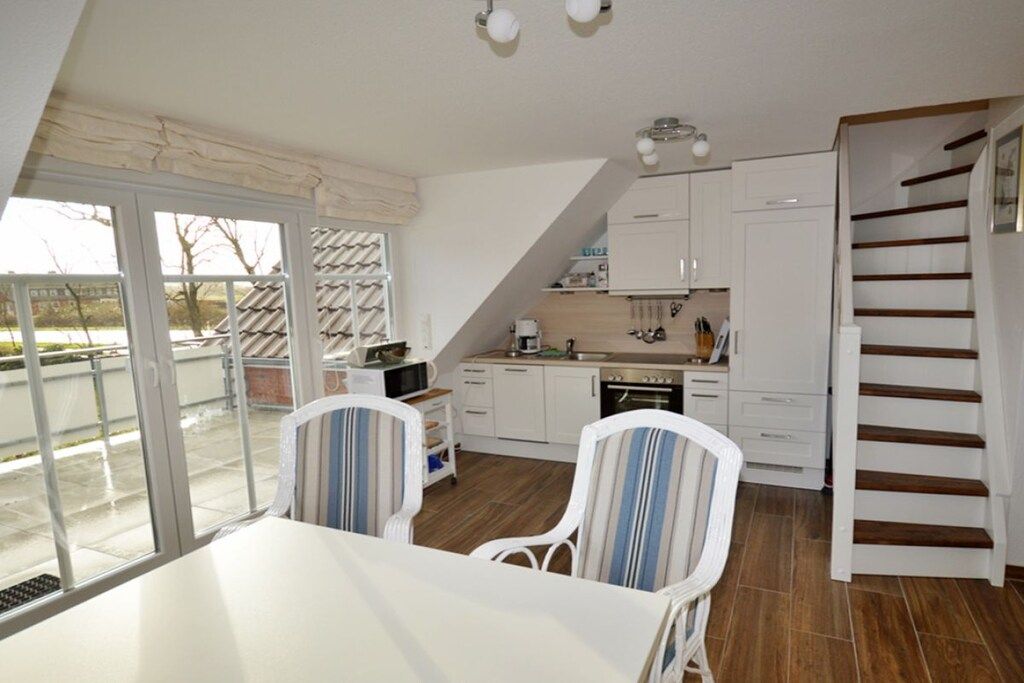 Une cuisine avec des meubles blancs, une table à manger avec des chaises et un escalier.