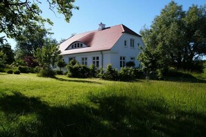 Une maison blanche avec un toit rouge, entourée d’un champ verdoyant et d’arbres.