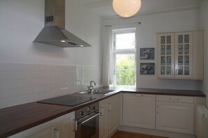 A kitchen with white cabinets, a stainless steel range hood, a window with curtains, and a view of greenery outside. - Holiday apartment with 3 bedrooms (Wohlenberg)