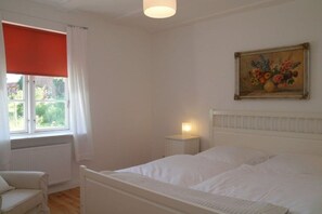 Une chambre à coucher avec un lit, une table de chevet, une chaise, une fenêtre avec des rideaux et un tableau au mur.
