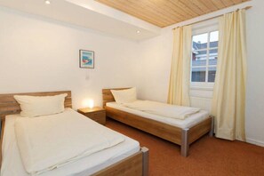 Ein Zimmer mit zwei Einzelbetten, einer Holzbalkendecke, einem Fenster mit Vorhängen und einem an der Wand hängenden Bild.