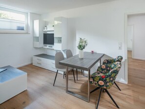 A modern kitchen with white cabinetry, a dining table, and a floral chair. - Nordhelmeck 6 Below (Norderney)