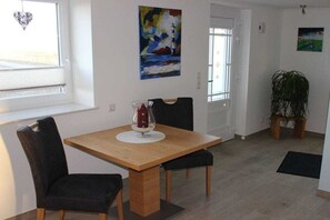 A dining area with a wooden table and four chairs, a vase with a red object, and a painting of a lighthouse on the wall. - Holiday home with 1 bedroom (Nordstrand)