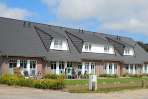 A two-story building with a flat roof, multiple windows, and a small outdoor seating area with chairs and a table. - Terraced house with 2 bedrooms (Sylt Ost\/ Tinnum)
