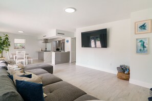 A modern living room with a grey sofa, a dining area with a white table and chairs, and a kitchen in the background. - Relaxation Time: Fenced yard, Screened Porch, Close to the Beach (Key Largo)