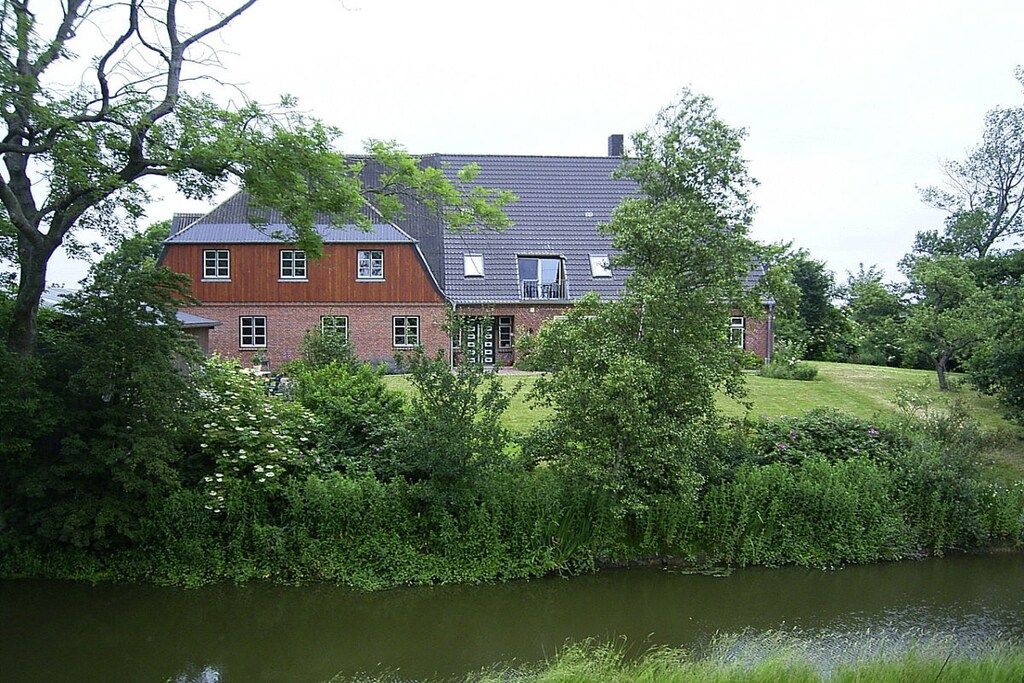 A large, two-story house with a brick facade and a grey roof, surrounded by lush greenery and a body of water.