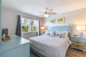 Ein Schlafzimmer mit einem Bett, einem Nachttisch, einem Schreibtisch, einem Deckenventilator und einem Fenster mit Vorhängen.