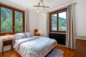 Ein Schlafzimmer mit einem großen Bett, zwei Nachttischen, einem Heizkörper und Blick auf einen Wald durch die Fenster.
