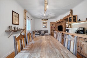 A dining room with a long wooden table, chairs, a chandelier, and a kitchen area with wooden cabinets and appliances. - Apartment 'Appartement de montagne' (Queige)