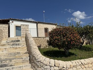 Ein Steinhaus mit einer Treppe, die zu einer Tür führt, ein kleiner Garten mit einem Baum und einem strahlend blauen Himmel.