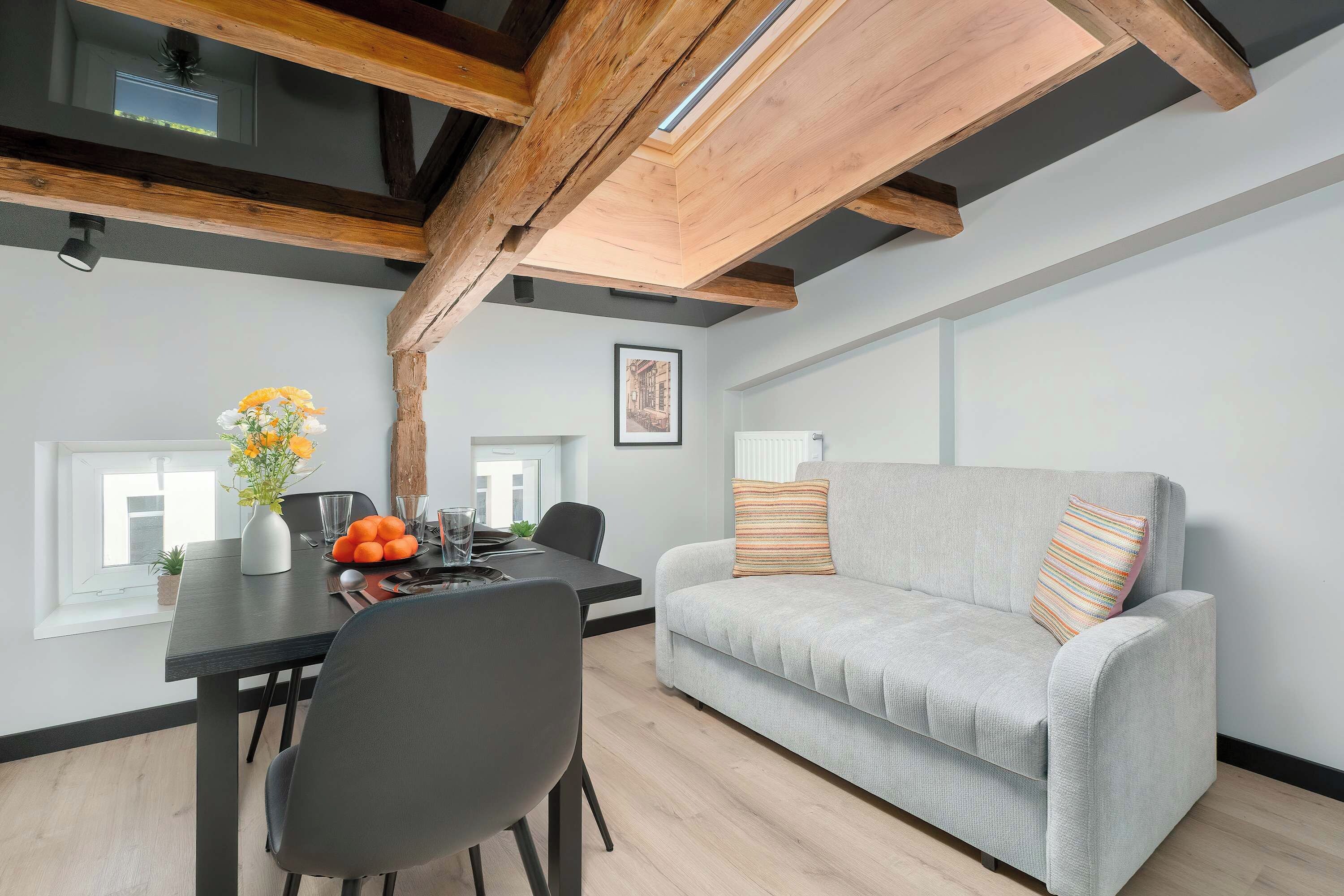 Un espace de vie moderne comprenant un coin repas, un canapé gris agrémenté de coussins à motifs et des poutres apparentes en bois.