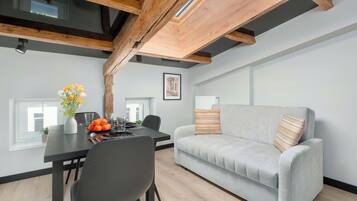 Un espace de vie moderne comprenant un coin repas, un canapé gris agrémenté de coussins à motifs et des poutres apparentes en bois.