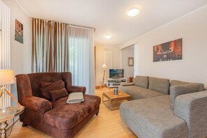 Ein Wohnzimmer mit braunem Sessel, grauem Sofa, Couchtisch aus Holz und einem Fernseher auf einem Lowboard.