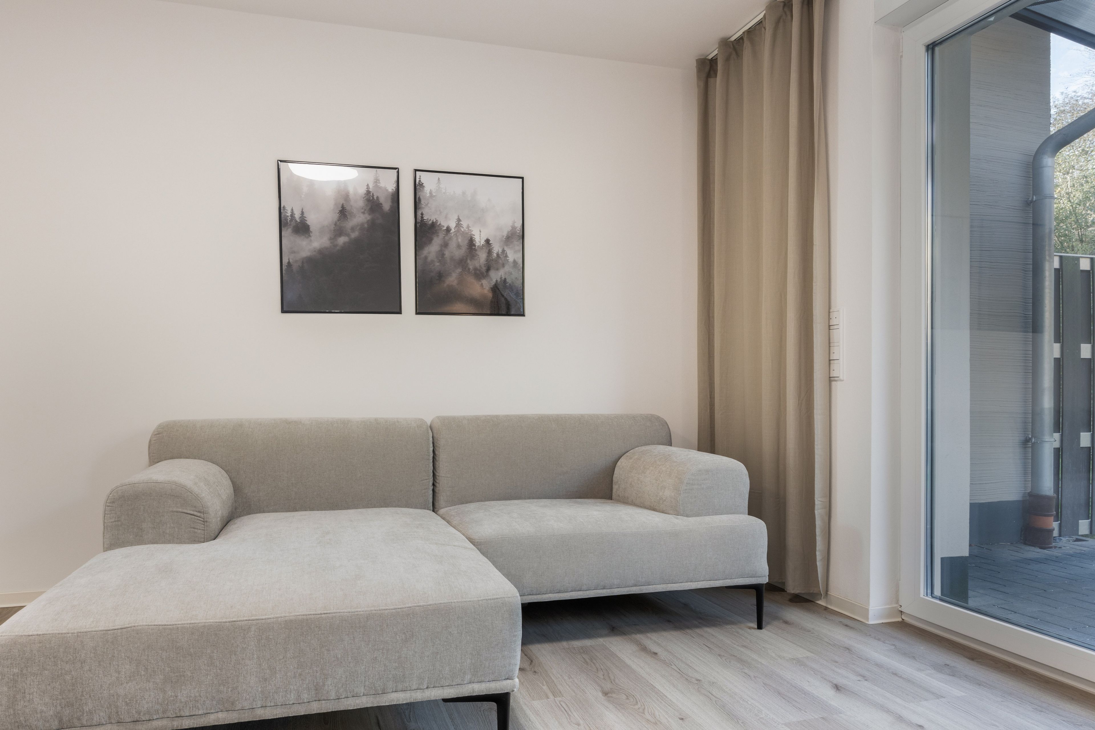 Un salon moderne avec un canapé d’angle gris, une porte vitrée donnant sur l’extérieur et deux tableaux encadrés accrochés au mur.