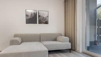 Un salon moderne avec un canapé d’angle gris, une porte vitrée donnant sur l’extérieur et deux tableaux encadrés accrochés au mur.