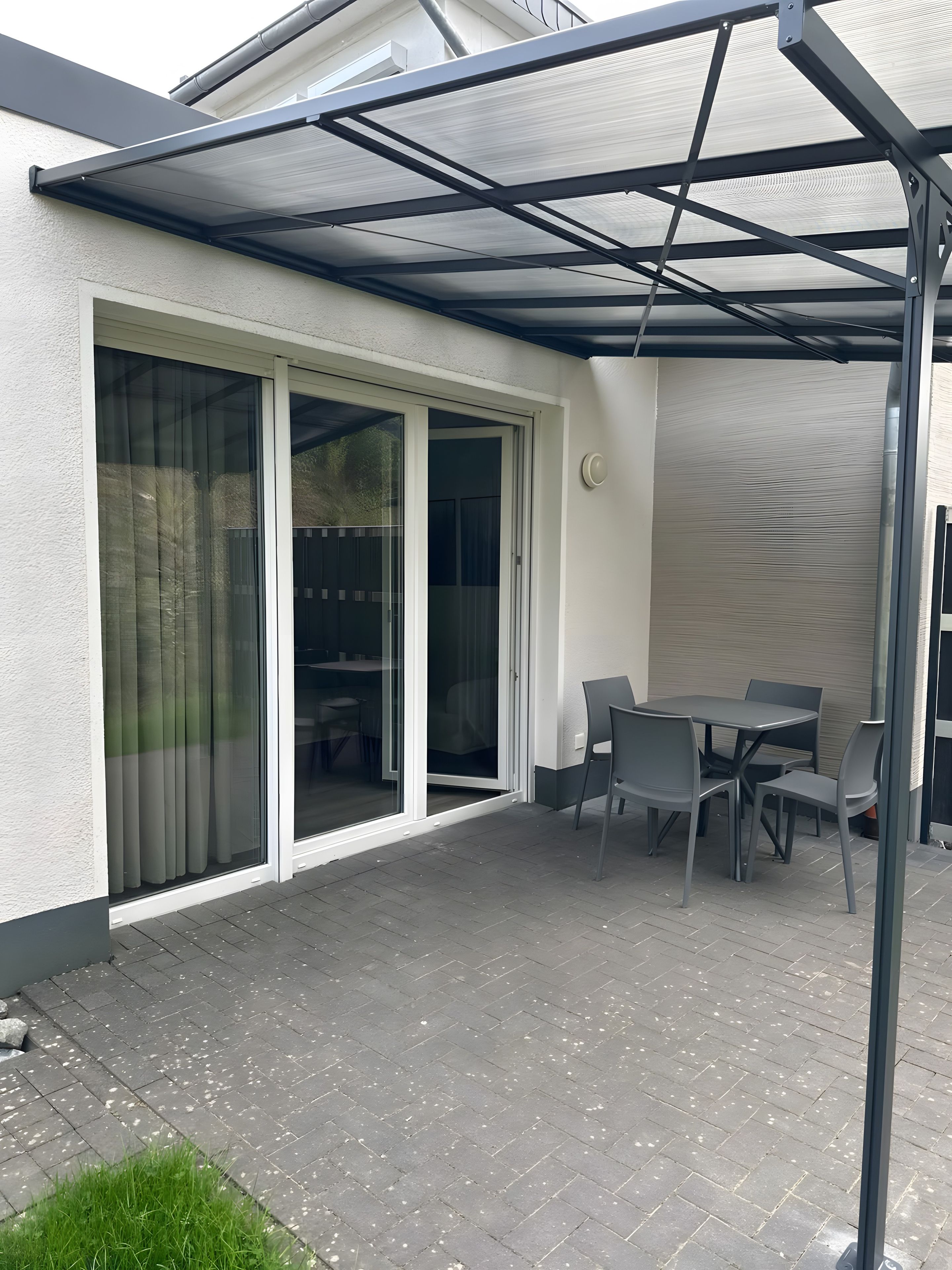 Ein überdachter Patio-Bereich mit Tisch und Stühlen, einer Schiebetür aus Glas und einer Metall-Dachkonstruktion.