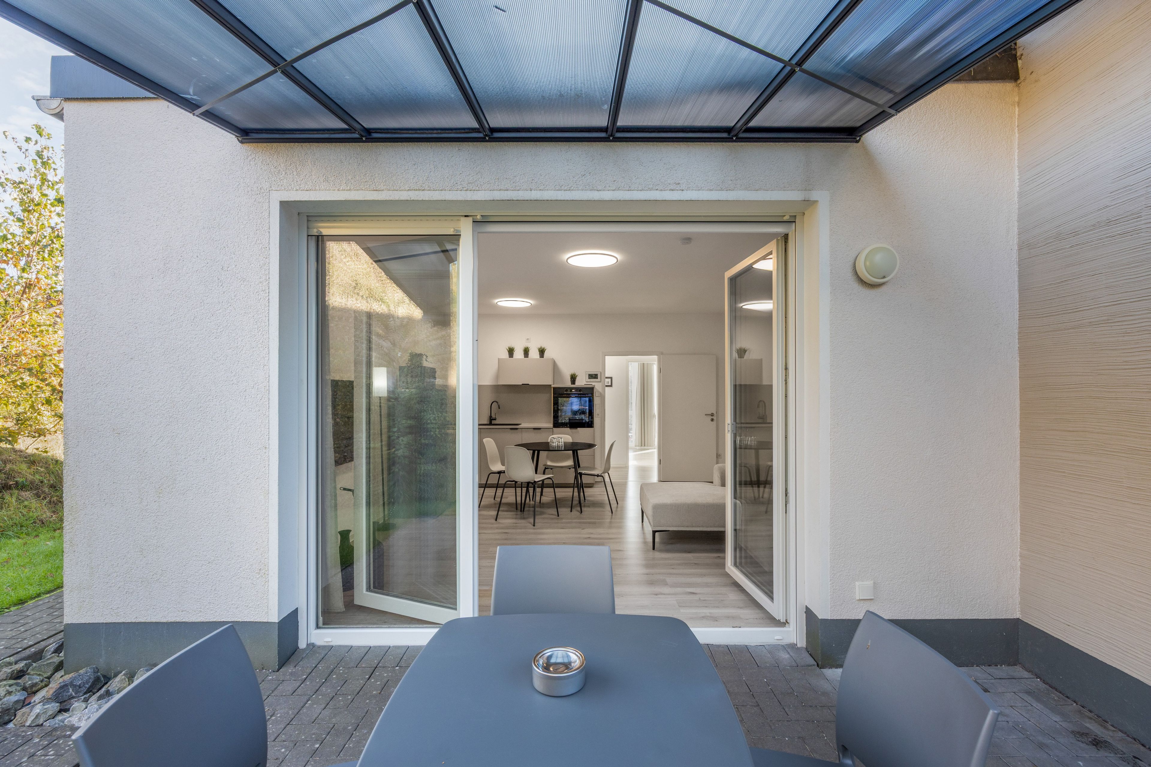 Eine Terrasse im Freien mit Tisch und Stühlen, eine Glastür, die zu einer Küche und einem Essbereich führt, und eine Decke mit Netzstruktur.