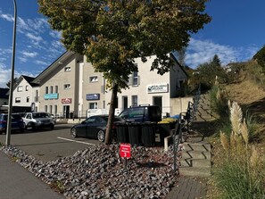 Ein Gebäude mit mehreren Schildern, darunter eines für eine Immobilienagentur und ein weiteres für ein Möbelgeschäft. Vor dem Gebäude befinden sich parkende Autos und ein Baum.