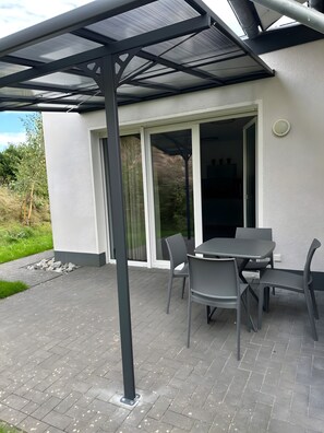 Eine Terrasse im Freien mit Tisch und Stühlen, ein überdachter Bereich mit Metallveranda und eine Glastür, die zu einem Innenraum führt.