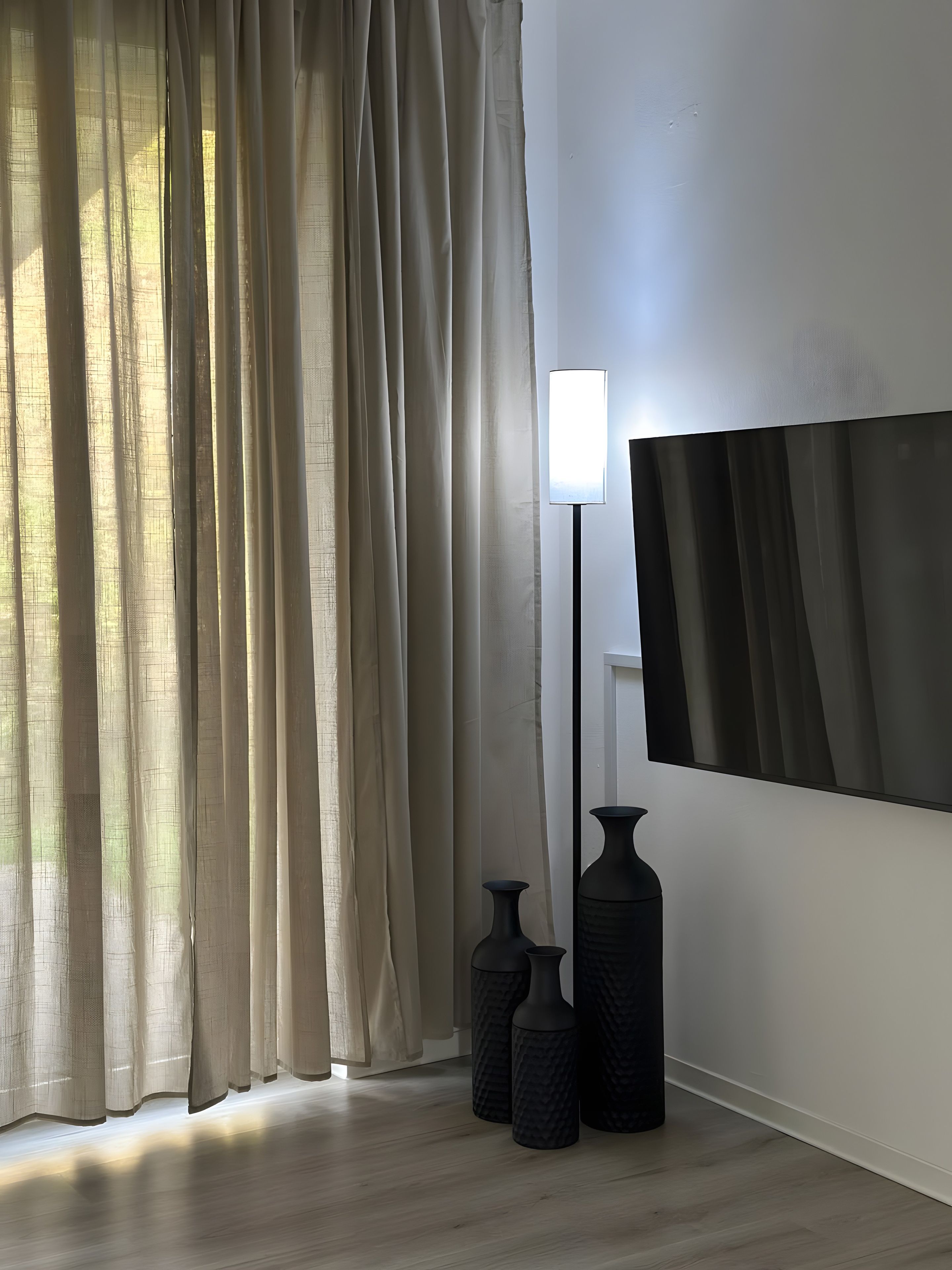 A room with a large window covered by sheer curtains, a floor lamp, and two black vases on the floor.