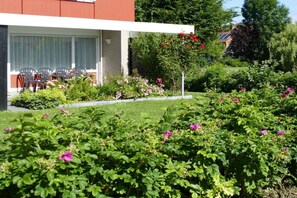 A modern building with a covered outdoor seating area, surrounded by well-maintained gardens with various flowering plants. - Holiday apartment Strandgut (Horumersiel)