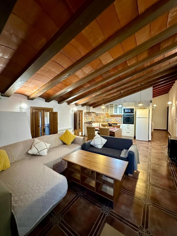 Un salón acogedor con techo alto, vigas de madera, un sofá, una mesa y una cocina al fondo.