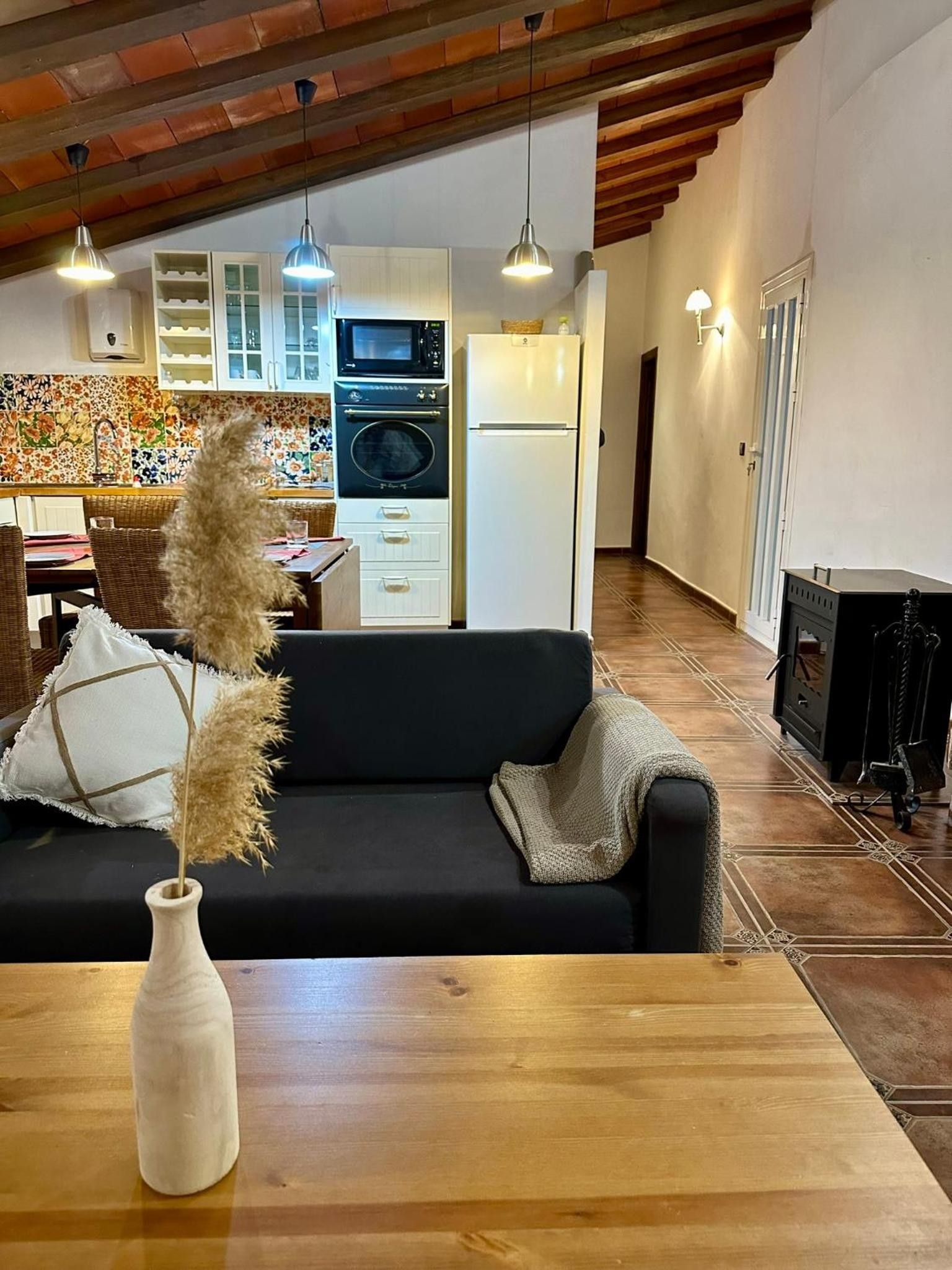 Een moderne woonkamer met een zwarte bank, een houten salontafel, een vaas met gedroogde planten, een keuken met witte kasten en apparatuur, en een open haard.