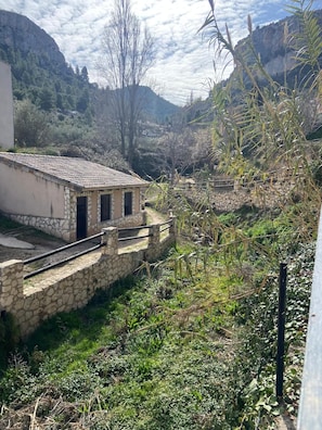 Un edificio de piedra con techo de tejas, rodeado de vegetación y montañas al fondo.