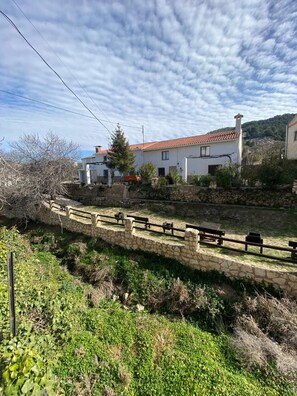 Un sendero de piedra conduce a un edificio blanco con tejado de tejas rojas, rodeado de vegetación y un cielo despejado.