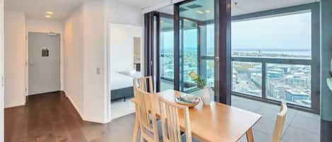 A modern hotel room with a large window offering a city view, a wooden dining table with chairs, and a small balcony area.
