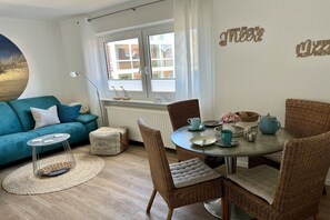 Ein gemütliches Wohnzimmer mit einem türkisfarbenen Sofa, Korbstühlen, einem runden Esstisch mit Tee-Service und einem großen Fenster mit Vorhängen.