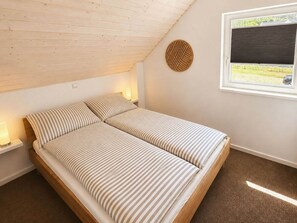 A bedroom with a sloped ceiling, a bed with striped bedding, bedside lamps, a window with a blind, and a wall-mounted circular object. - Holiday Home in Nordhorn With Swimming Pool (Nordhorn)