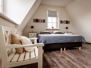 Ein Schlafzimmer mit schrägem Dach, einem Bett mit blauer Bettdecke, einer weißen Bank mit Kissen und zwei Nachttischen mit Lampen.
